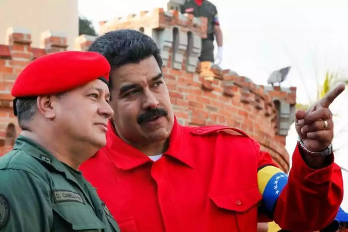 Dos hombres con uniforme militar y chaqueta roja conversan frente a una estructura de ladrillos.