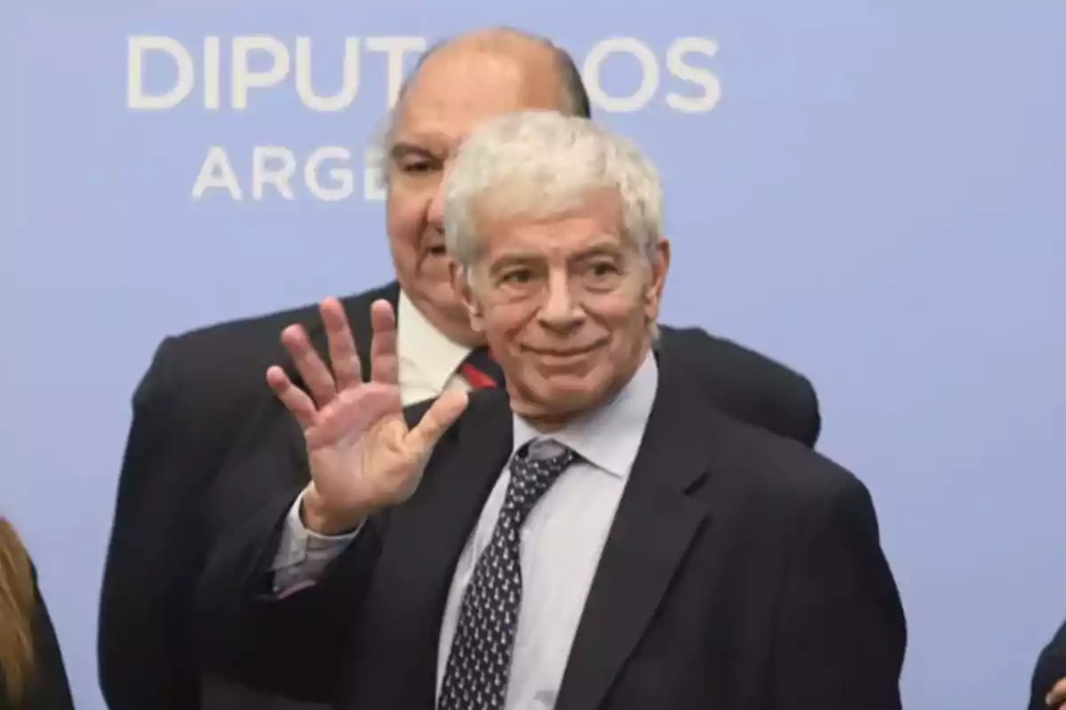 Un hombre de cabello canoso y traje oscuro saluda con la mano levantada frente a un fondo azul que dice "Diputados Argentina".