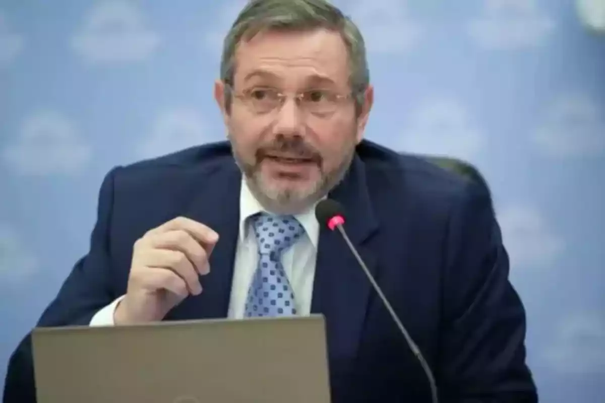 Hombre con barba y gafas hablando en un micrófono durante una conferencia, vestido con traje y corbata, frente a una computadora portátil.