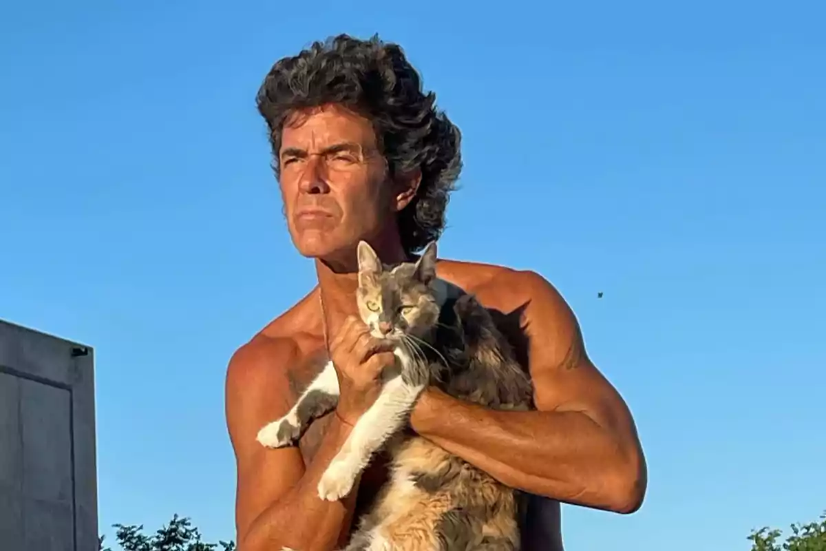 Un hombre sin camisa sostiene a un gato mientras mira hacia el horizonte bajo un cielo azul.