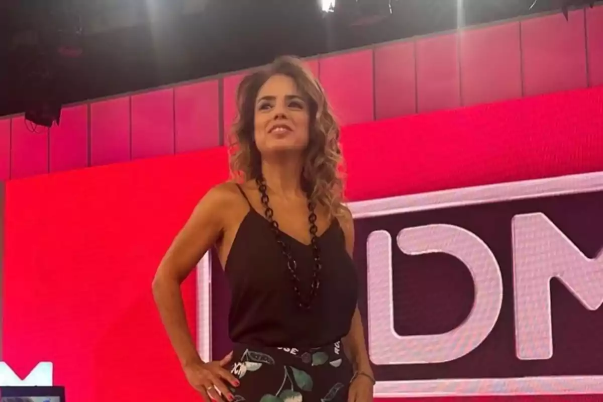 Mujer de pie en un set de televisión con fondo rojo y letras grandes.