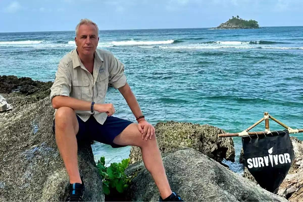 Un hombre sentado en una roca junto al mar con una bandera que dice "Survivor" y una isla al fondo.