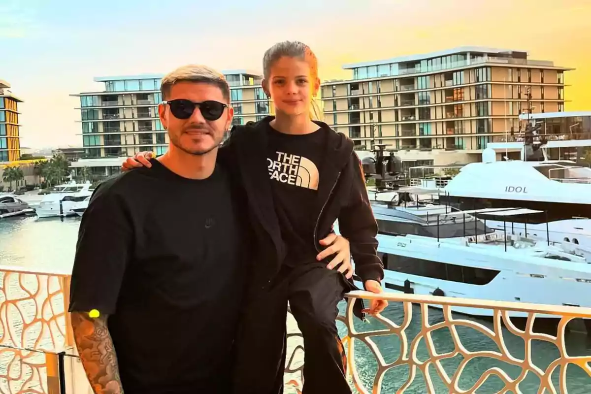 Un hombre con gafas de sol y una niña posan juntos en un balcón con vista a un puerto deportivo al atardecer.