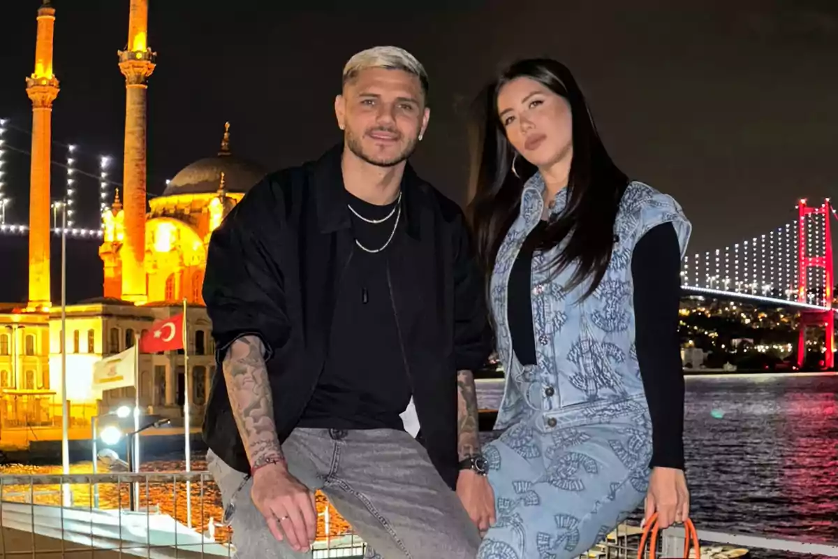 A couple posing in front of a nighttime landscape with an illuminated mosque and a bridge in the background.