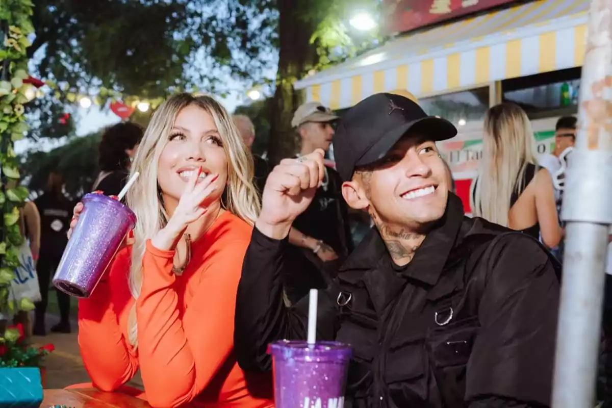 Two smiling people sitting at an outdoor table, a woman in an orange dress holding a shiny glass and a man in a black cap also holding a similar glass, with a background of lights and greenery.