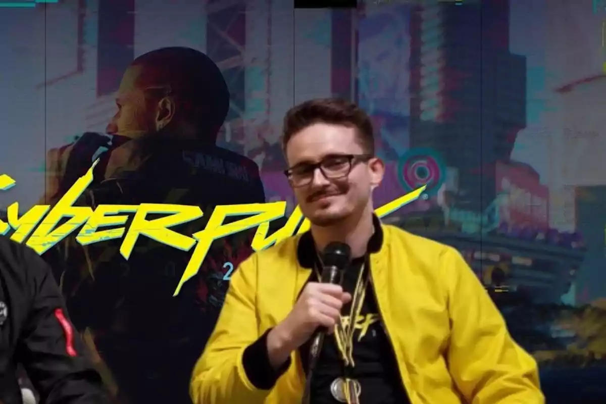 Un hombre con gafas y chaqueta amarilla sostiene un micrófono frente a un fondo con el logo de Cyberpunk 2077.