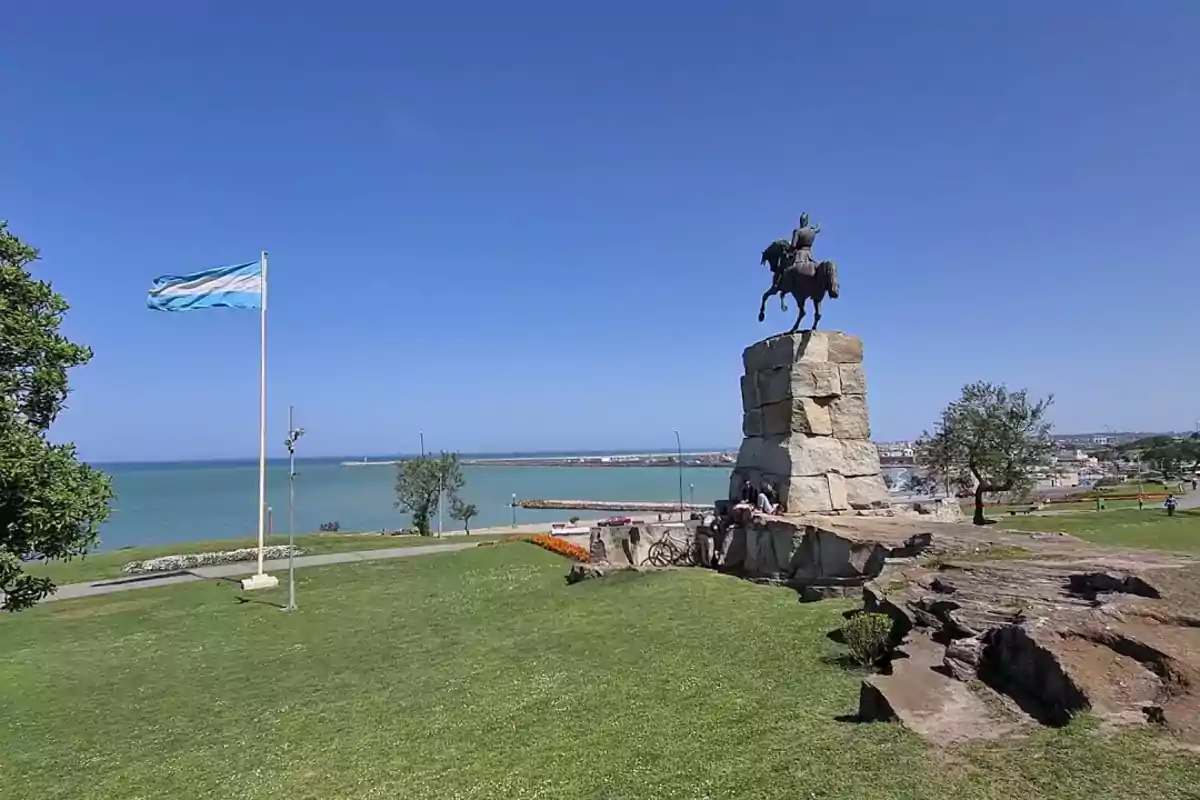 Una estatua ecuestre sobre un pedestal de piedra con una bandera ondeando cerca y el mar de fondo en un día soleado.