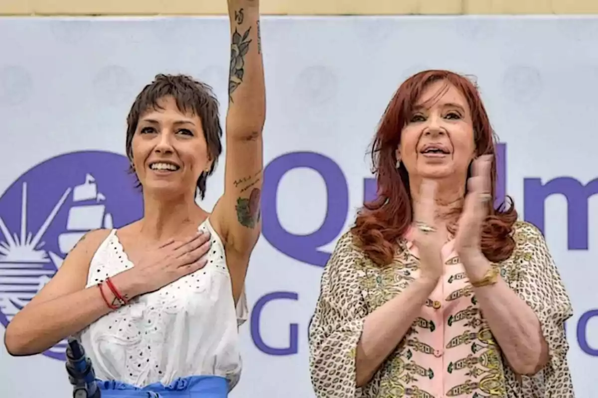 Dos mujeres en un evento público, una con el brazo levantado y la otra aplaudiendo, con un fondo que tiene un logotipo y texto parcialmente visible.