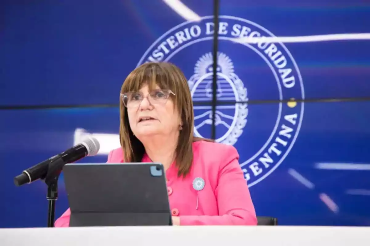 Una persona con gafas y chaqueta rosa habla en un micrófono frente a un fondo azul con un emblema oficial.