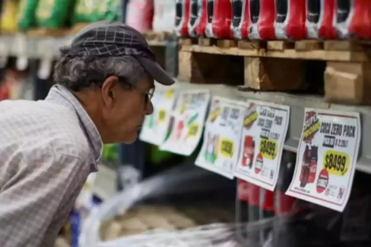 Un hombre con gorra observa de cerca las etiquetas de precios en una tienda, donde se exhiben botellas de refresco en un estante.