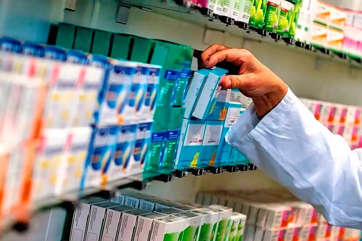 Una persona con bata blanca selecciona una caja de medicamentos de un estante lleno de productos farmacéuticos.