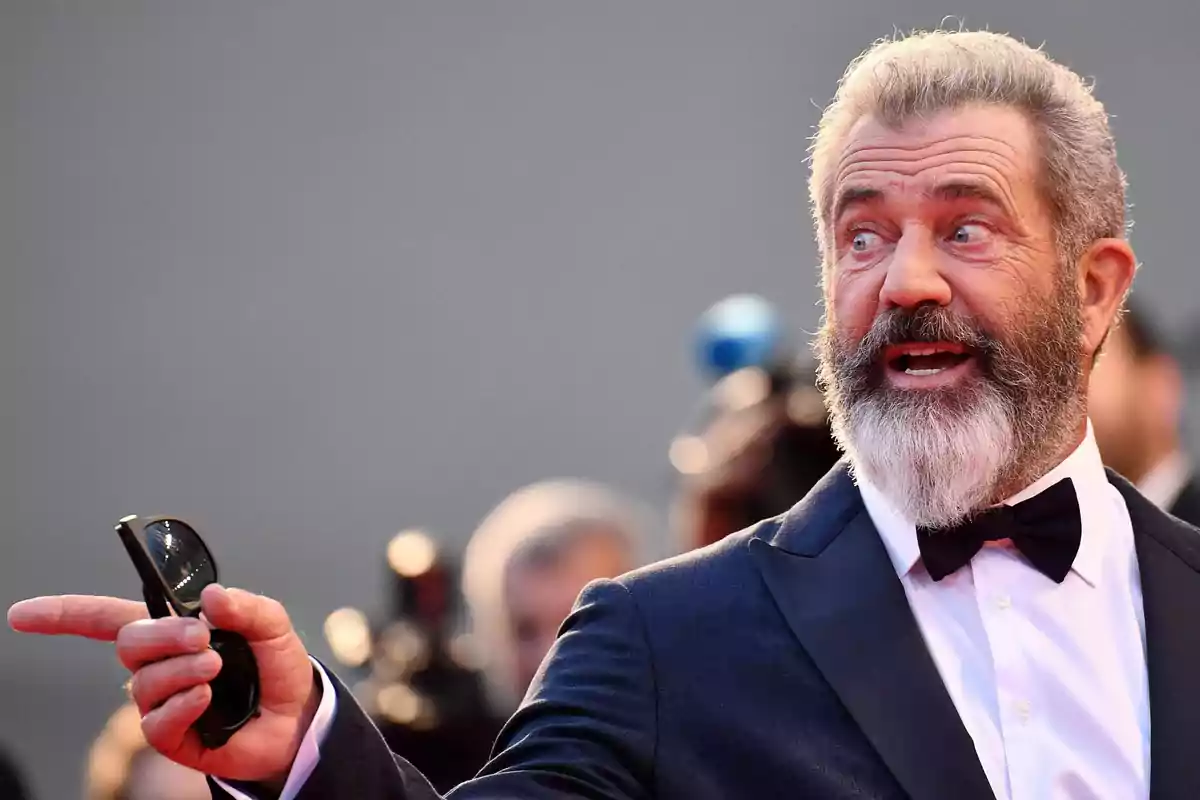 Un hombre con barba y traje oscuro señala con la mano mientras sostiene unas gafas de sol.