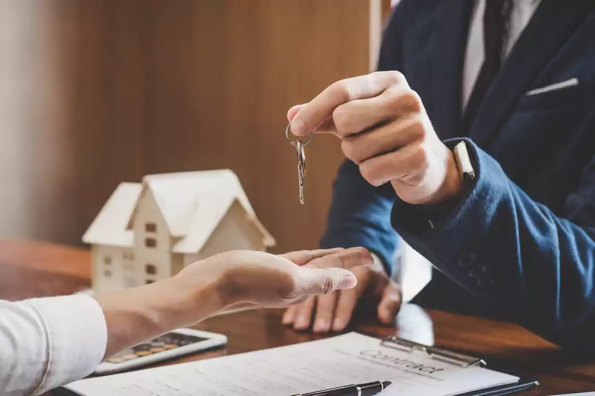 Una persona entrega una llave a otra sobre una mesa con un contrato y una maqueta de casa.