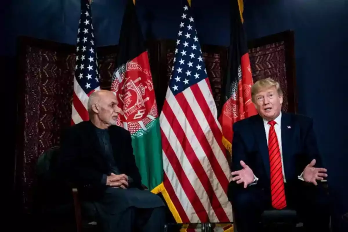 Dos hombres sentados conversando frente a banderas de Estados Unidos y Afganistán.