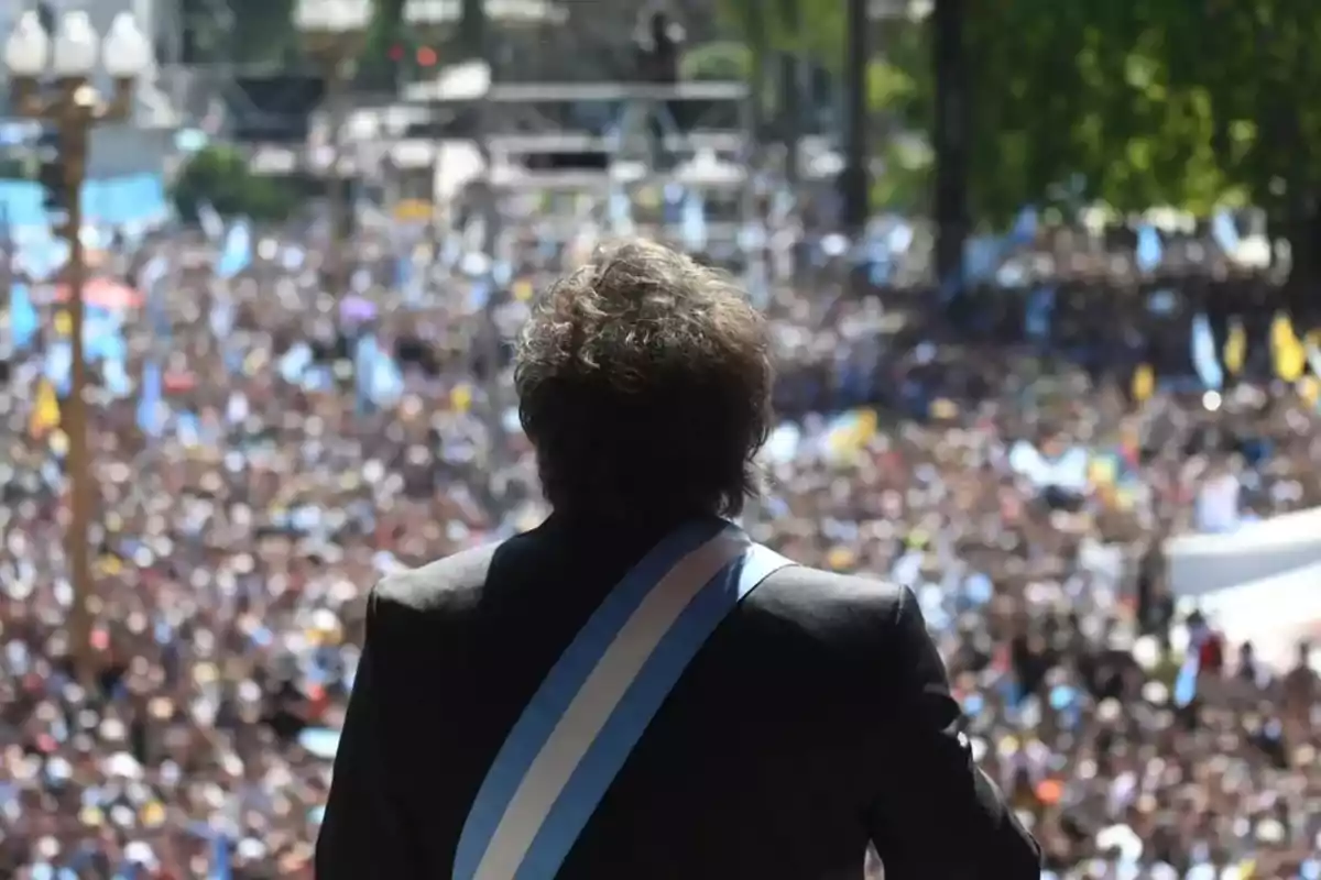 Una persona con una banda presidencial de espaldas frente a una multitud.