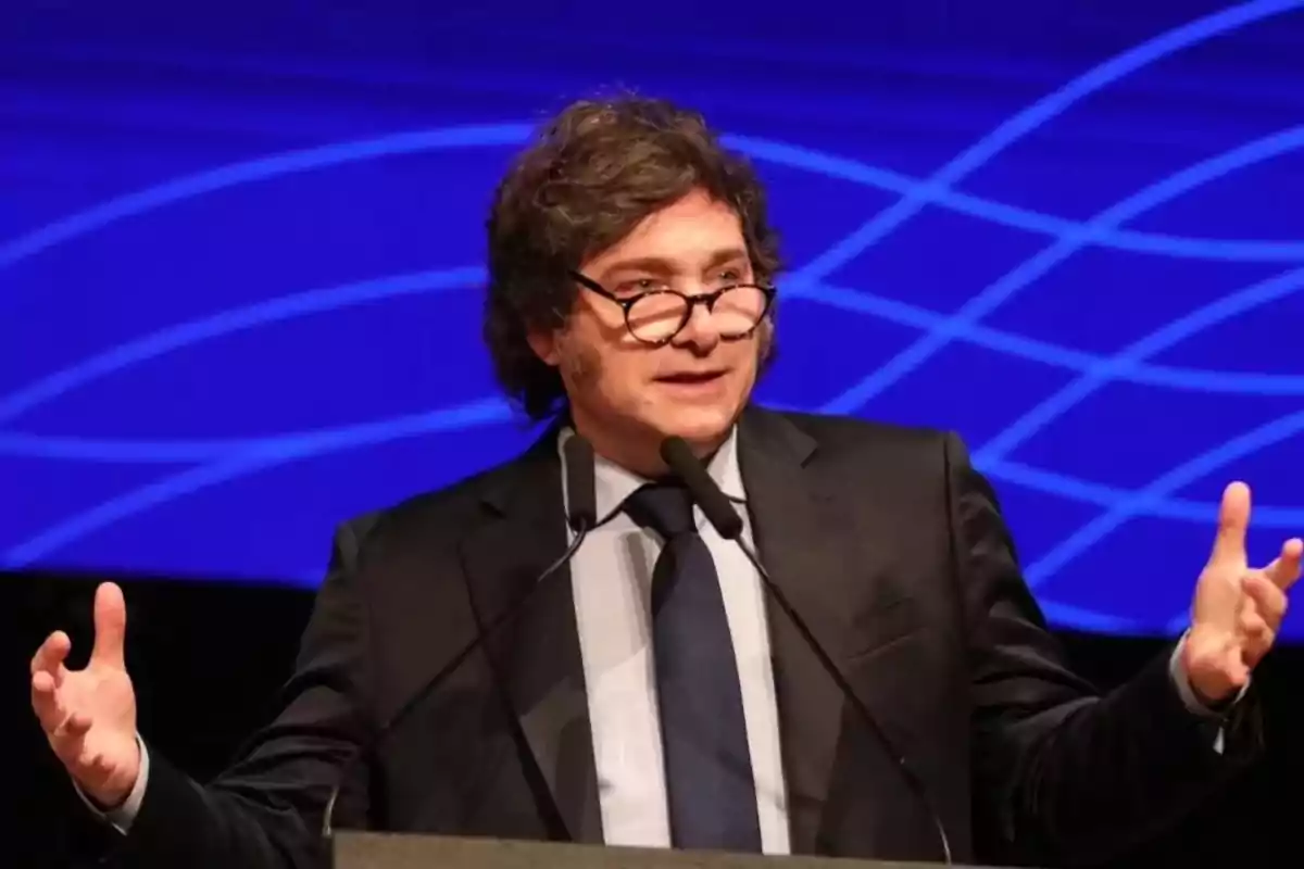 Un hombre con gafas y traje hablando en un escenario con fondo azul.