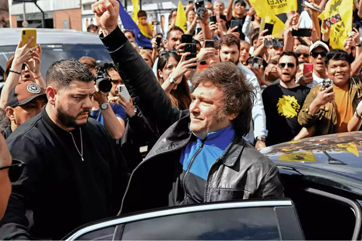 Un hombre con chaqueta de cuero levanta el puño mientras es rodeado por una multitud de personas que lo fotografían y graban con sus teléfonos móviles.