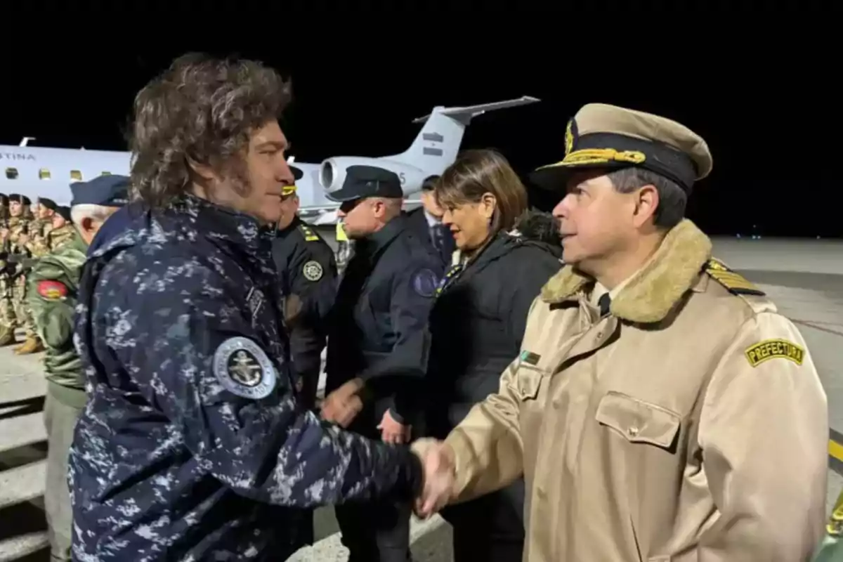 Dos hombres uniformados se dan la mano en un aeropuerto, con un avión y varias personas uniformadas en el fondo.