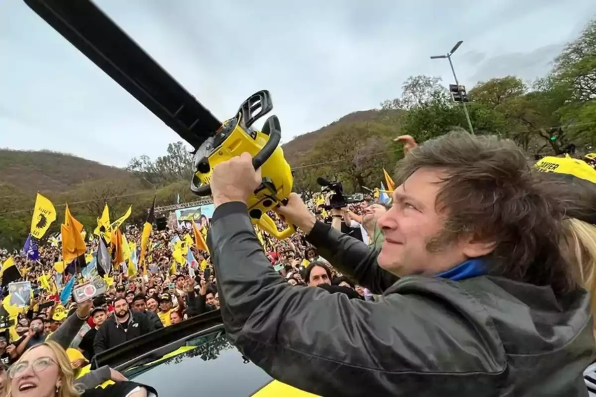 Una persona sostiene una motosierra amarilla en un evento al aire libre con una multitud de personas y banderas de colores.