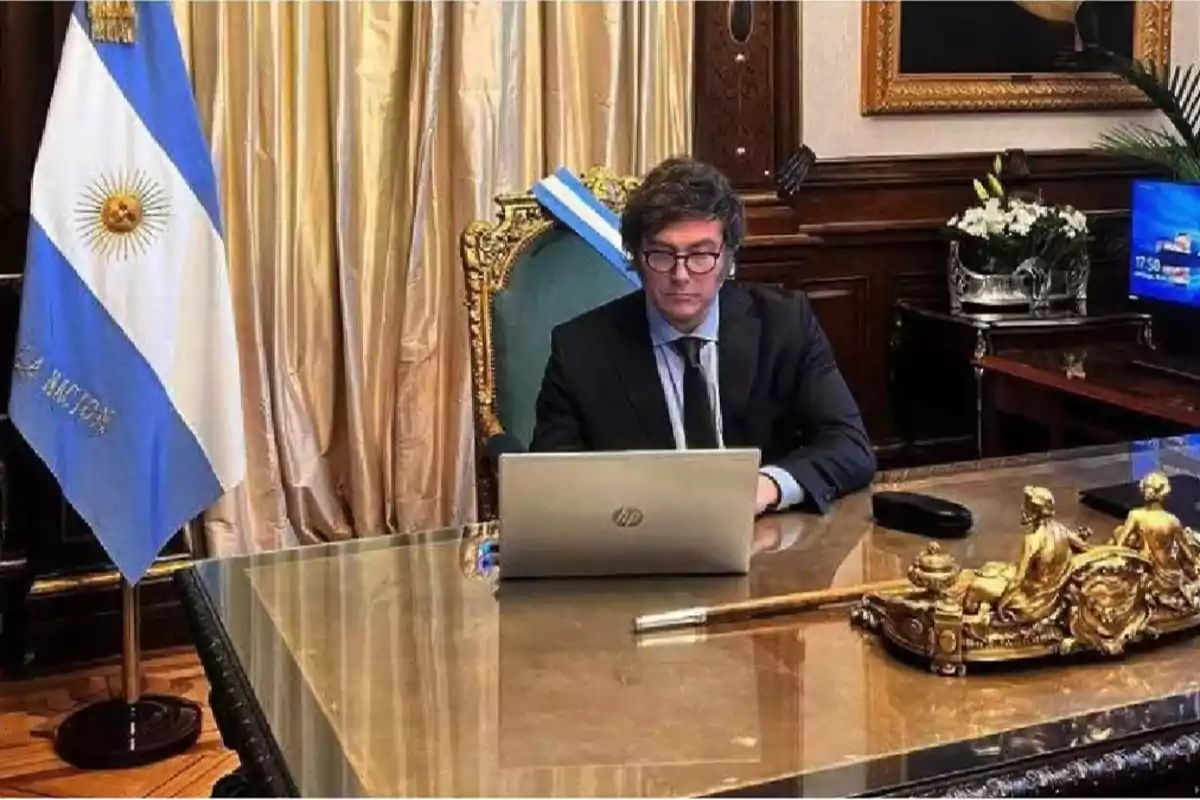 Un hombre con traje y corbata trabaja en una computadora portátil en un escritorio elegante, con una bandera de Argentina a su lado y un bastón dorado decorativo sobre la mesa.