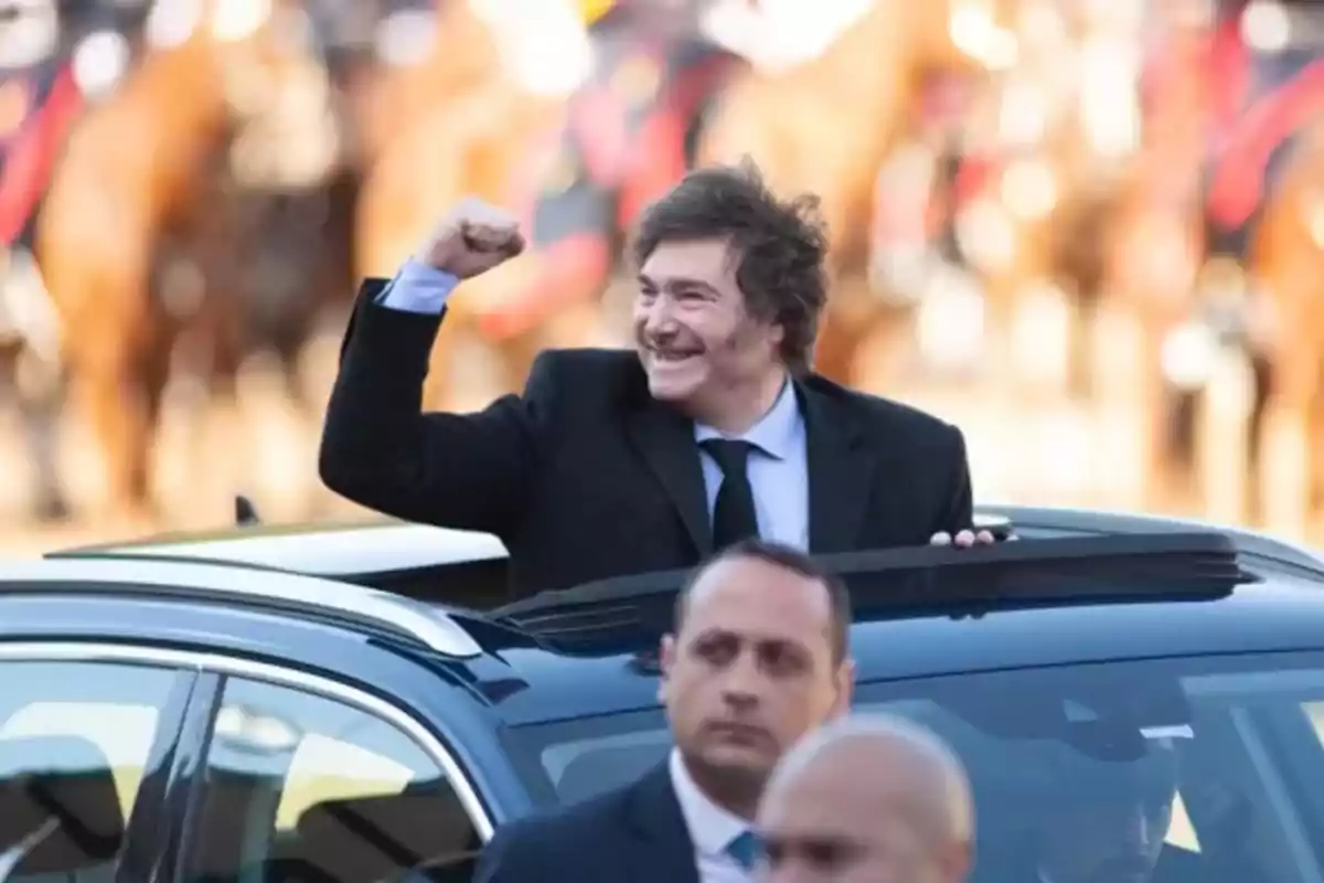 Un hombre sonriente con traje oscuro saluda desde el techo de un automóvil rodeado de personas y caballos desenfocados al fondo.