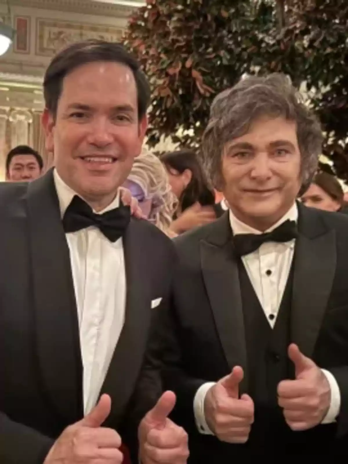 Dos hombres vestidos de traje y corbata de moño posan sonriendo con los pulgares hacia arriba en un evento elegante.