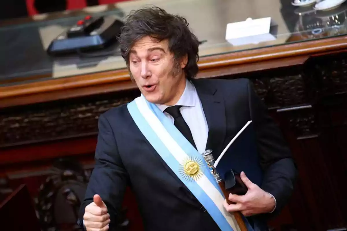 Un hombre con traje y banda presidencial gesticula con entusiasmo en un entorno formal.