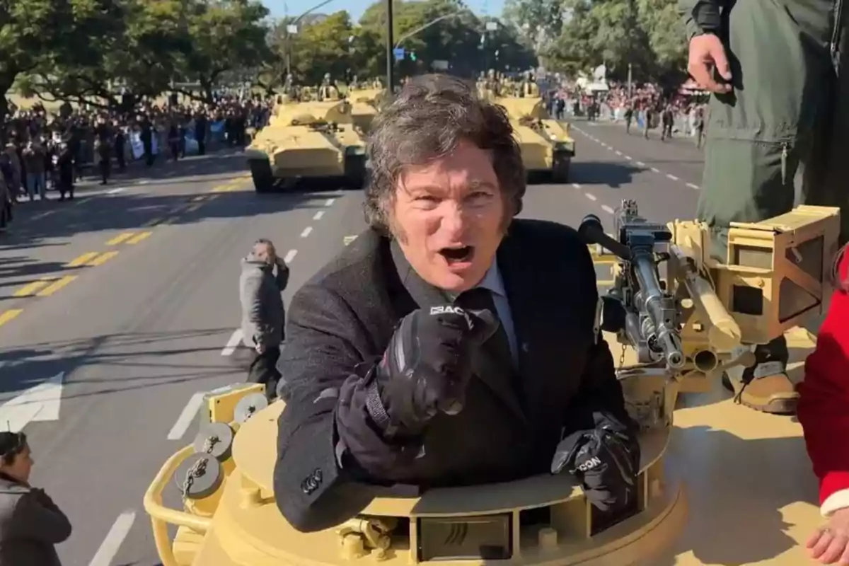 Una persona con guantes negros está asomada en un vehículo militar durante un desfile, rodeada de tanques y una multitud observando en la calle.