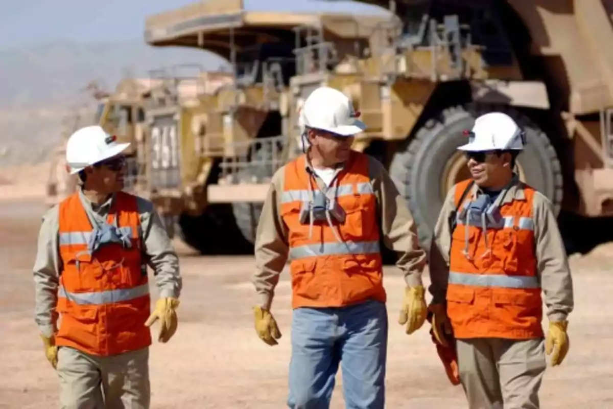 Tres trabajadores con cascos y chalecos de seguridad naranjas caminando en un sitio de construcción con maquinaria pesada en el fondo.