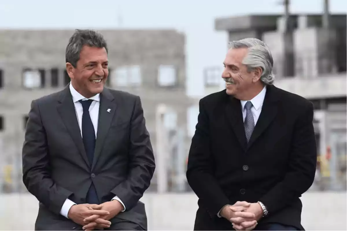 Dos hombres vestidos de traje sonríen mientras están sentados uno al lado del otro en un entorno urbano.