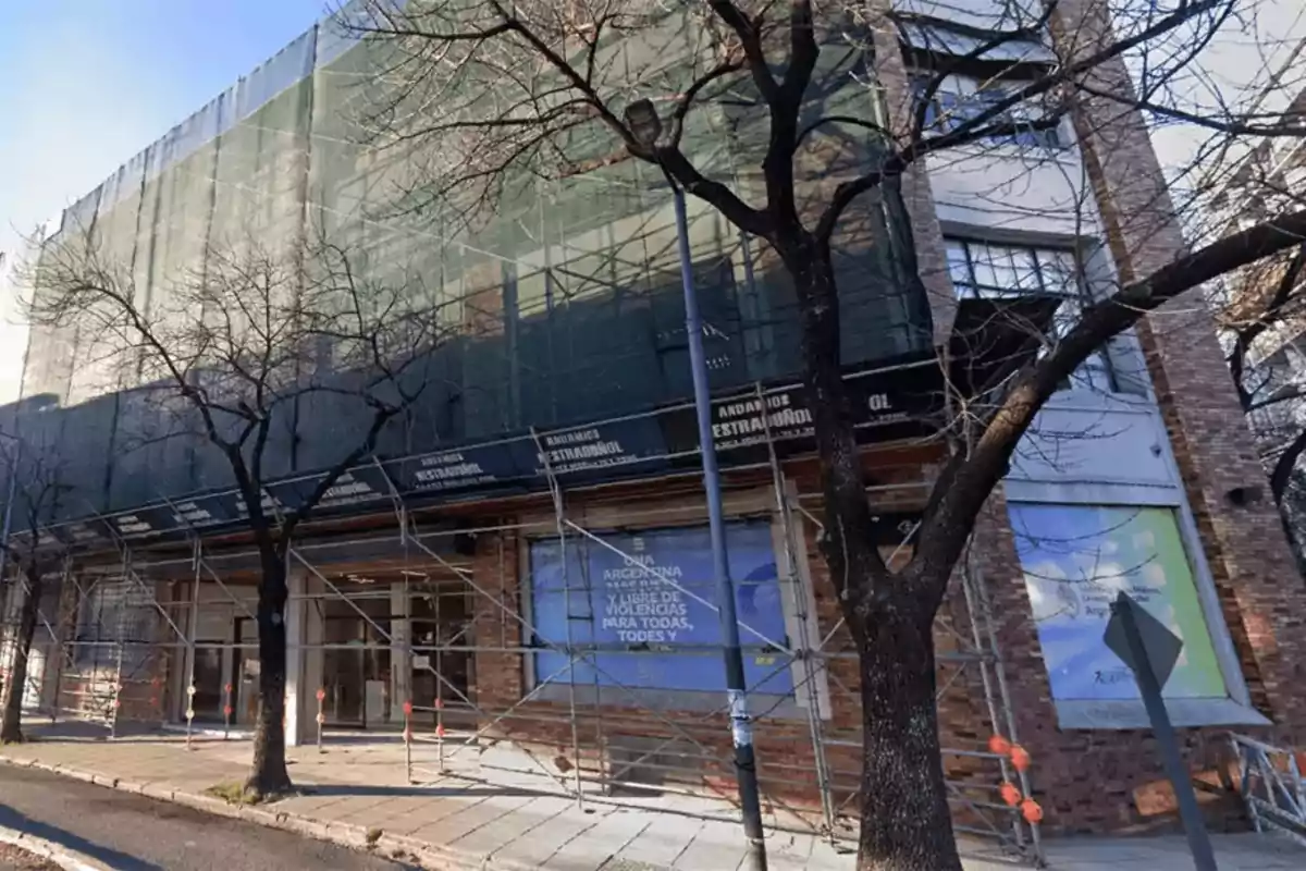 Edificio en construcción con andamios y una malla verde cubriendo la fachada, árboles sin hojas en la acera y un cartel que menciona la lucha contra las violencias.