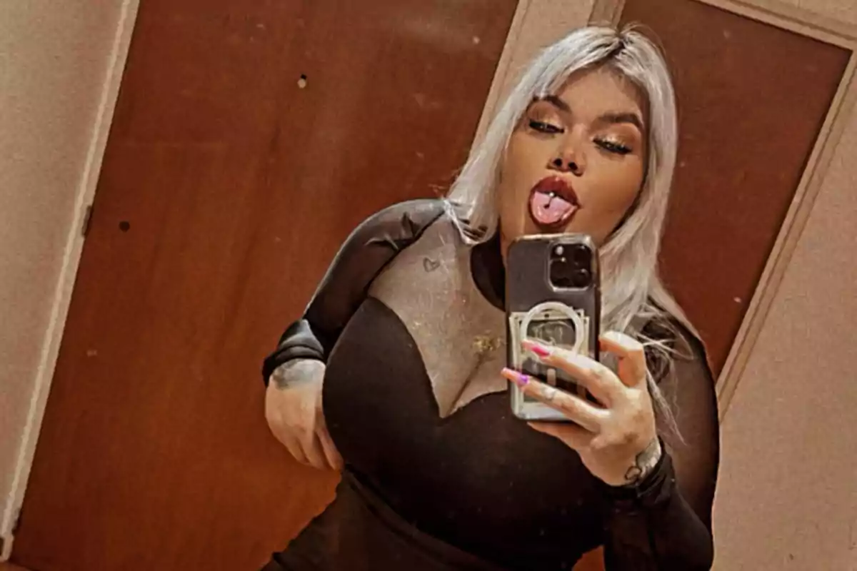 A person with blonde hair and striking makeup takes a selfie in front of a mirror, sticking out their tongue and showing a piercing, wearing a black dress with sheer panels.