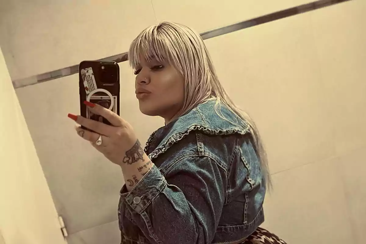 A person with blonde hair and a denim jacket takes a selfie in front of a mirror.