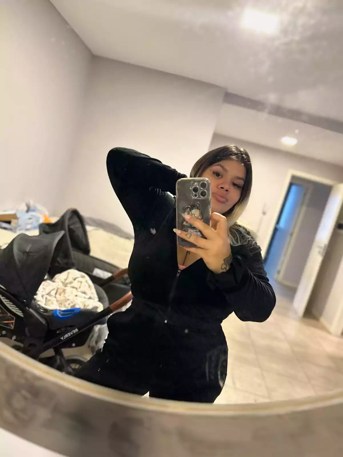 A person taking a selfie in front of a mirror in a room with a baby stroller in the background.