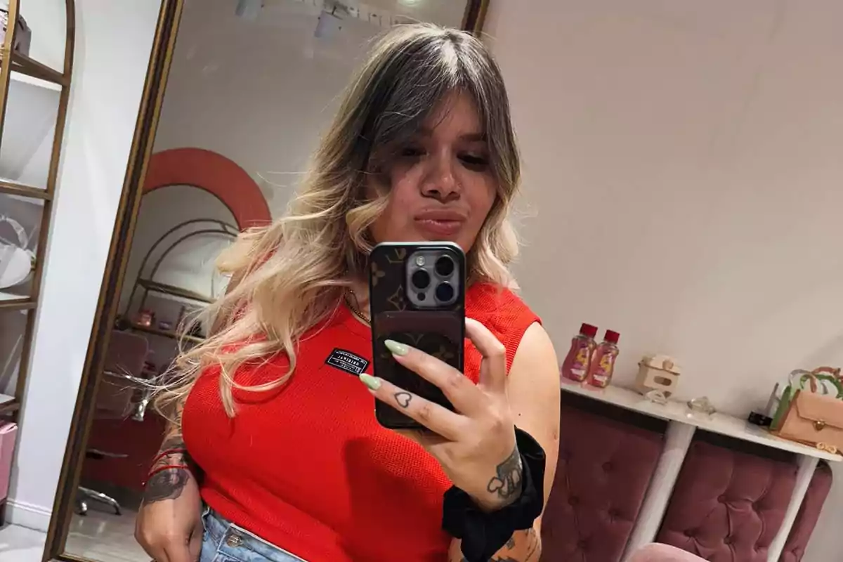 A person with blonde and dark hair takes a selfie in front of a mirror, wearing a red t-jersey and holding a mobile phone.