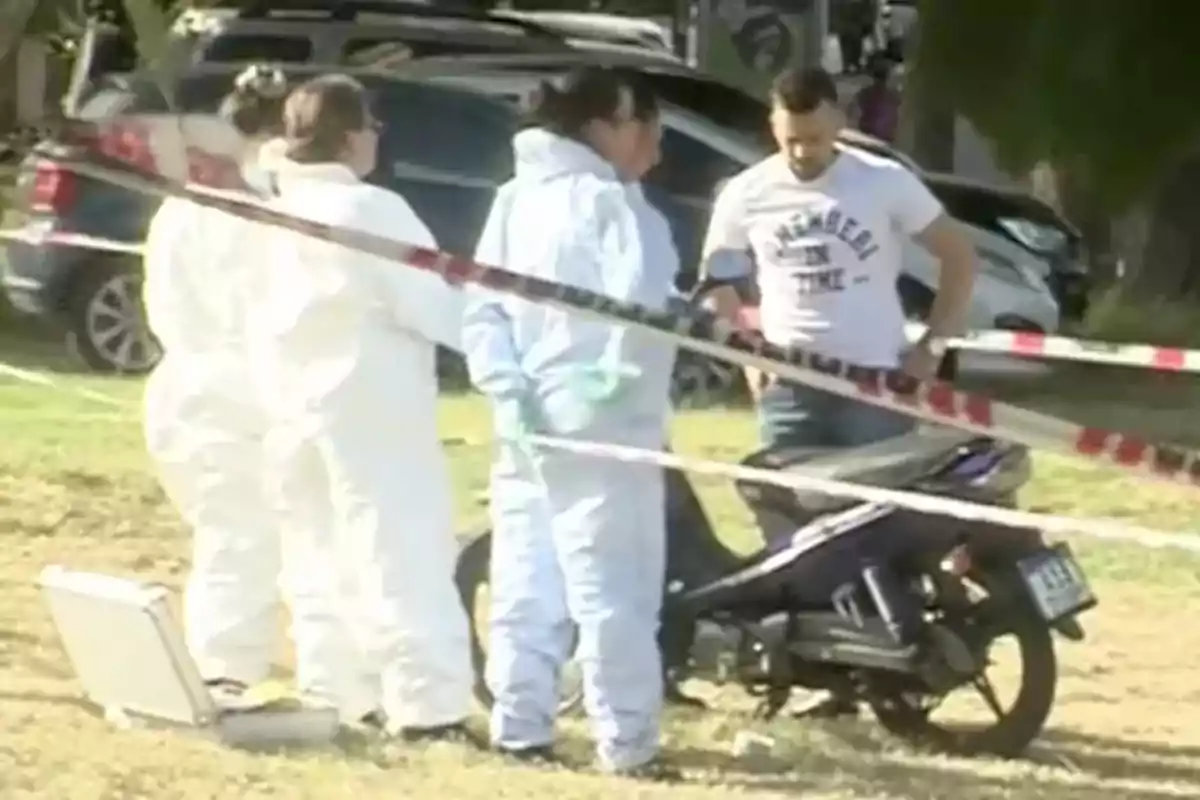 Personas con trajes de protección y un hombre de pie junto a una motocicleta en un área acordonada.