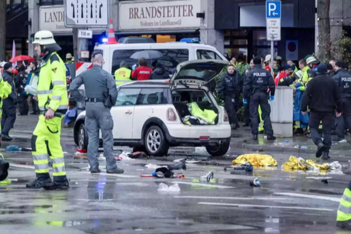 A group of police officers and emergency personnel surround a white car with the trunk open on a street where there is debris and emergency blankets scattered.