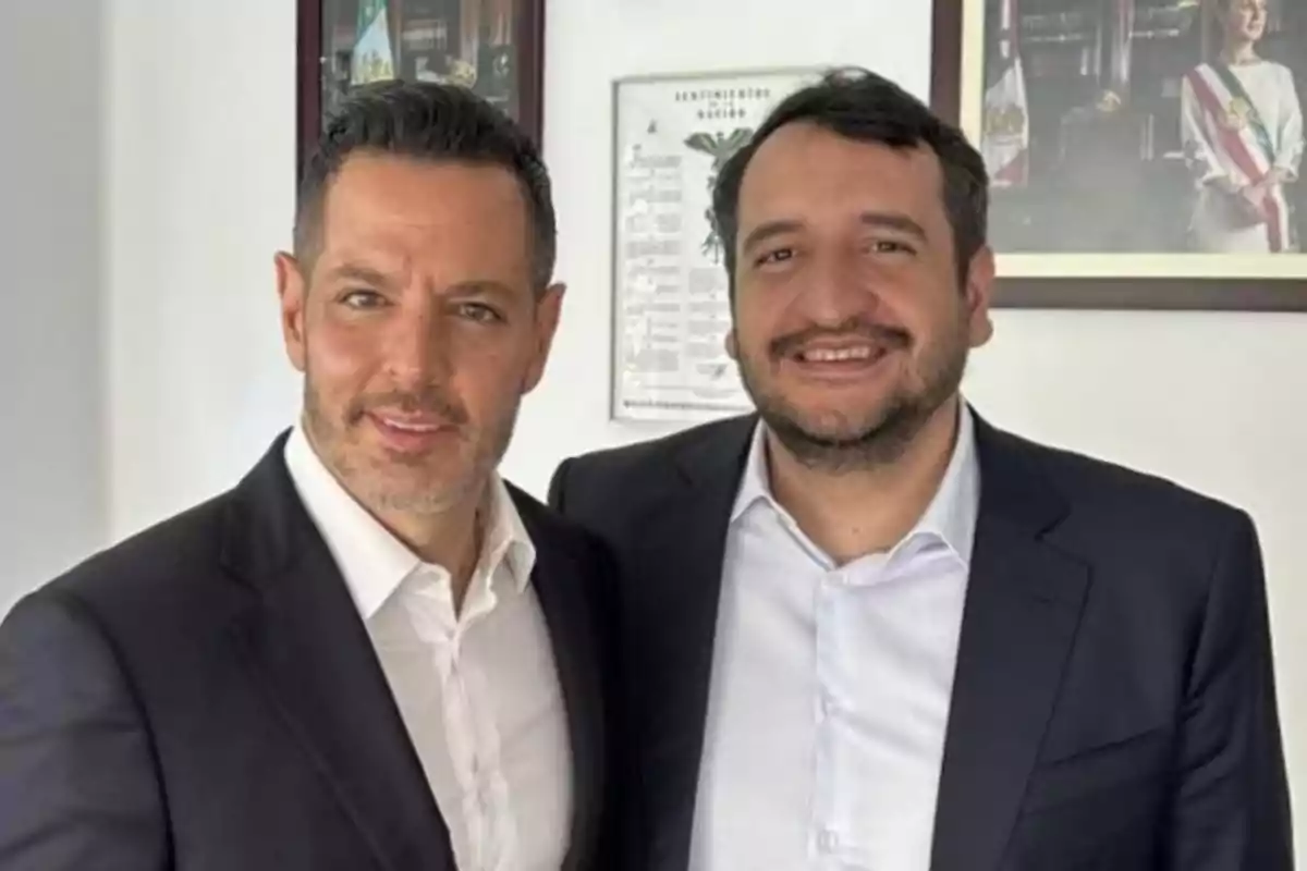 Two men dressed in dark suits pose together in an office with paintings on the wall.