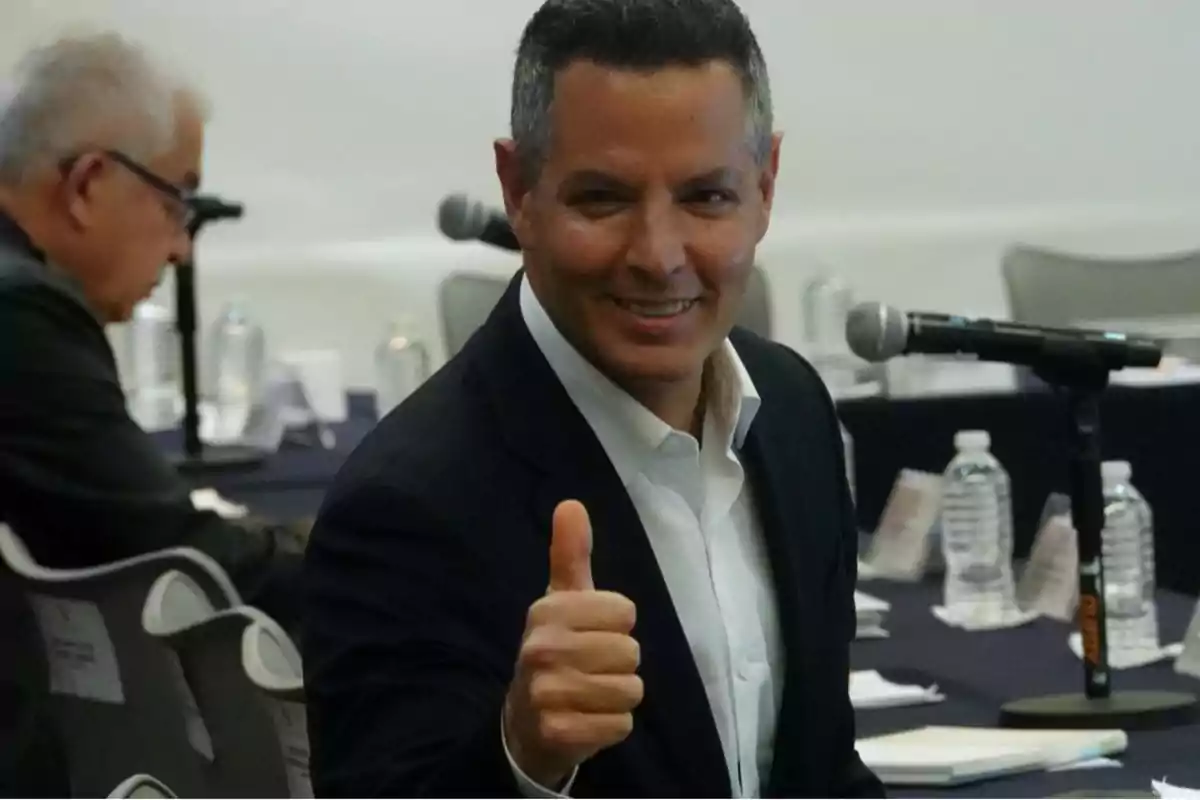 A smiling man in a dark suit gives a thumbs-up in a conference room.