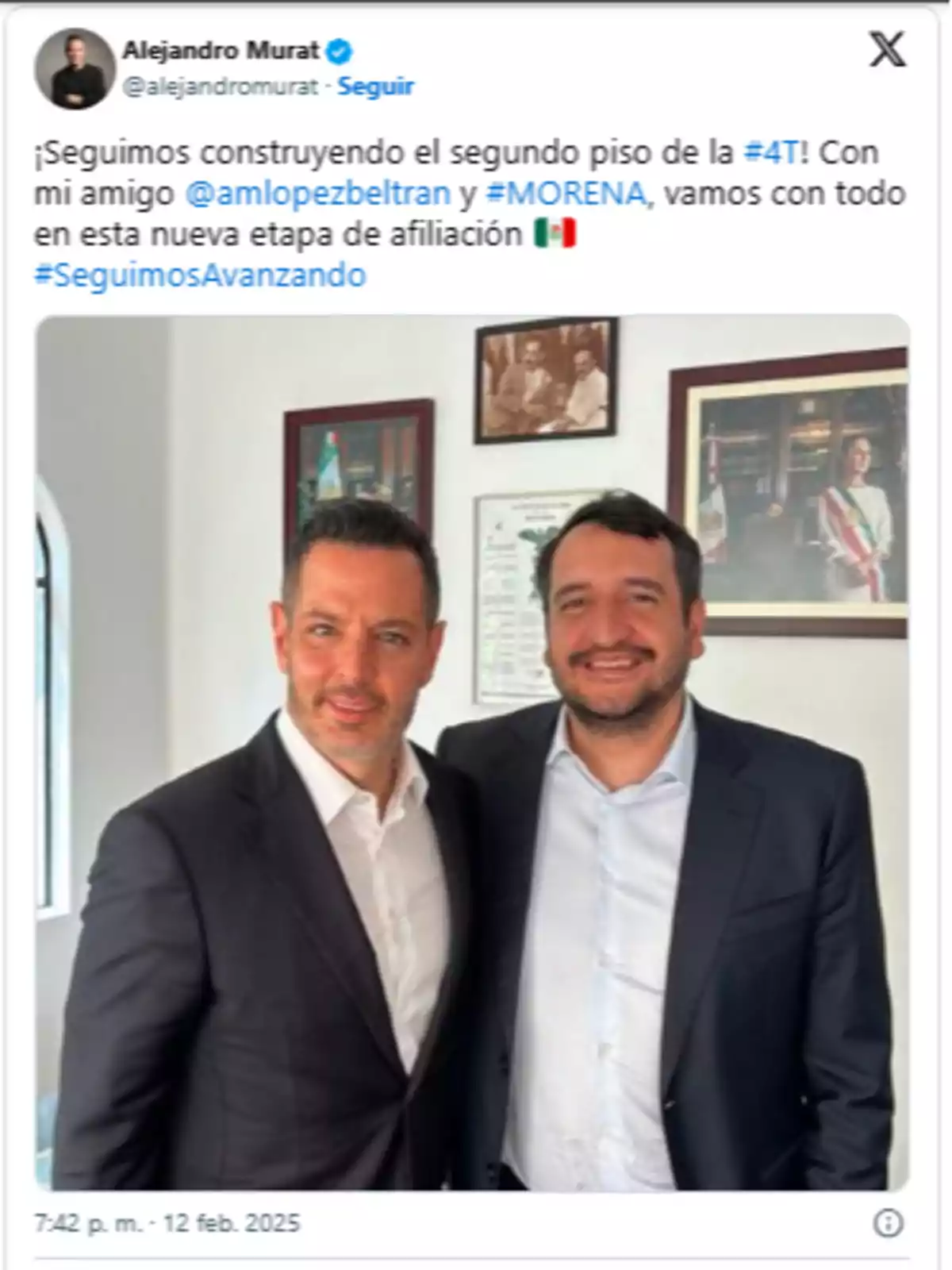 Two men dressed in dark suits pose together smiling in an office with paintings on the wall.