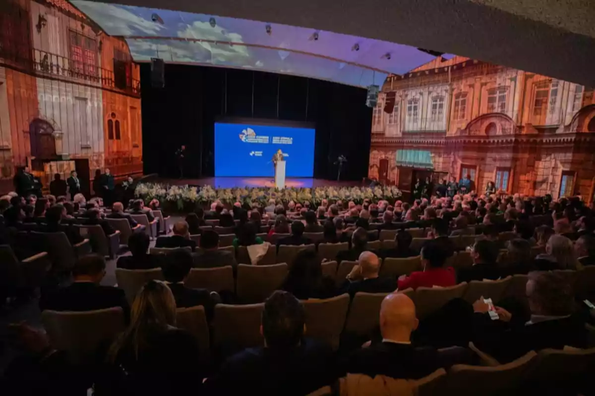 Una conferencia en un auditorio lleno de personas con un escenario decorado con proyecciones de edificios históricos y una pantalla azul al fondo.