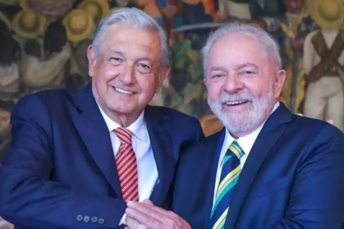 Dos hombres mayores sonrientes vestidos con trajes formales se dan la mano frente a un mural colorido.