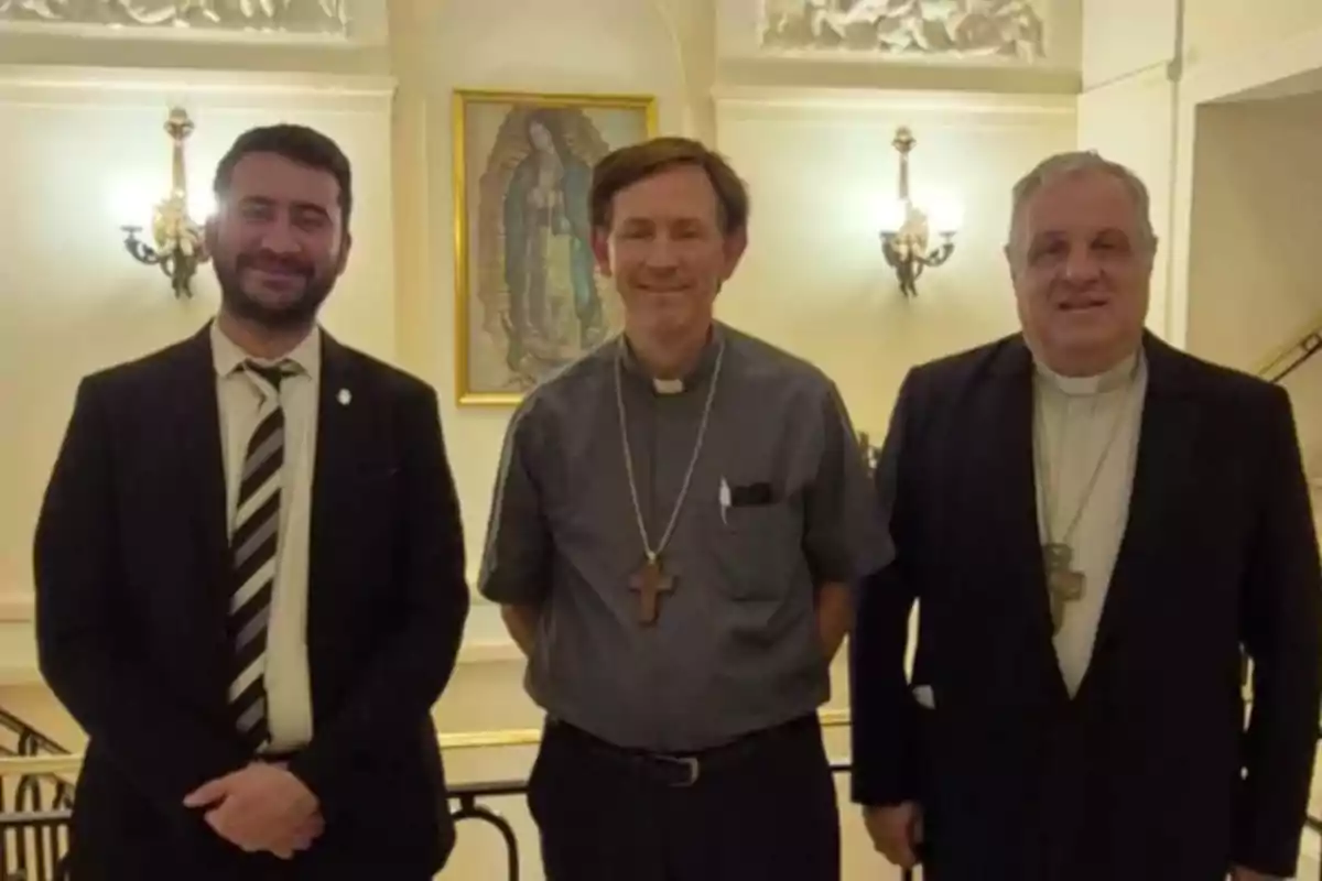 Tres hombres posan juntos en un interior elegante con una imagen religiosa en la pared detrás de ellos.