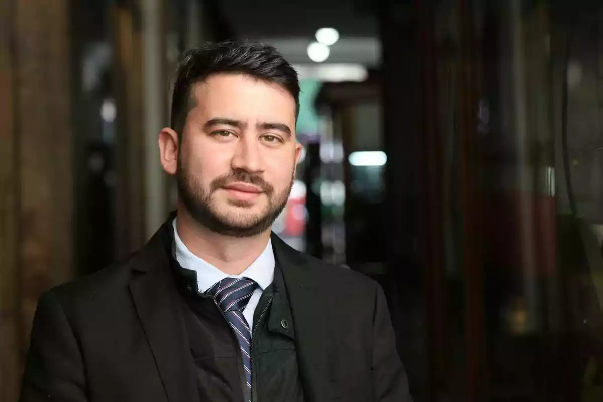 Hombre con barba y cabello oscuro vistiendo traje y corbata en un entorno urbano.