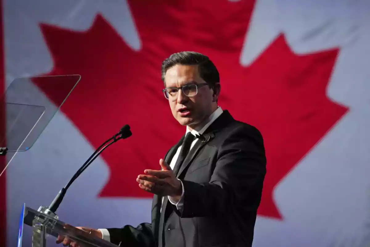 Un hombre en traje habla en un podio frente a una bandera de Canadá.