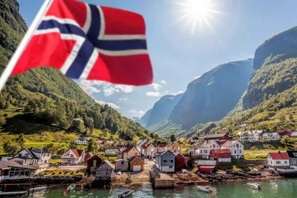 Paisaje de un pueblo noruego con montañas al fondo y una bandera de Noruega ondeando en primer plano bajo un cielo soleado.