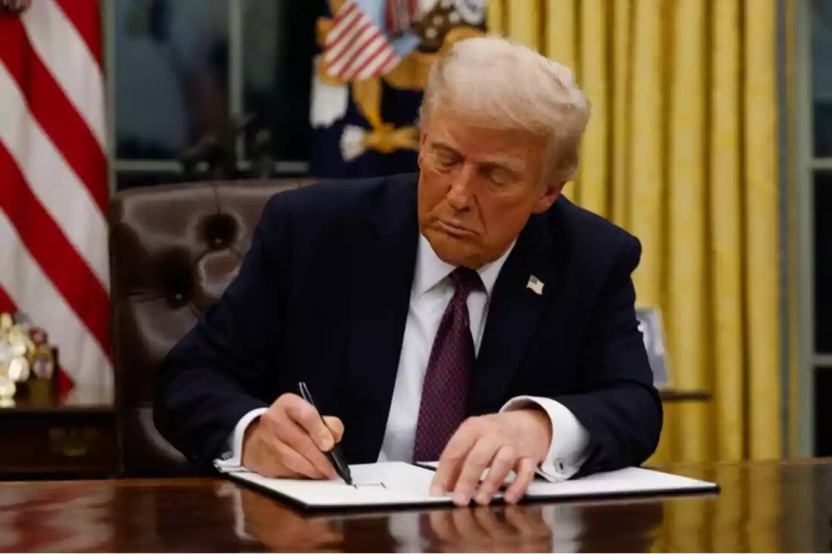 Un hombre de traje oscuro firmando un documento en un escritorio con una bandera estadounidense y cortinas doradas de fondo.