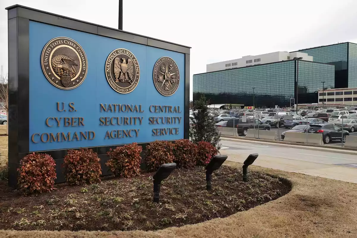 Un cartel azul con los emblemas de U.S. Cyber Command, National Security Agency y Central Security Service frente a un edificio de oficinas y un estacionamiento.