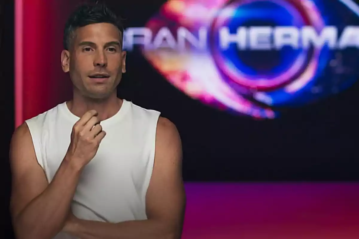 Un hombre con camiseta sin mangas blanca está de pie frente a un fondo con el logo de un programa de televisión.