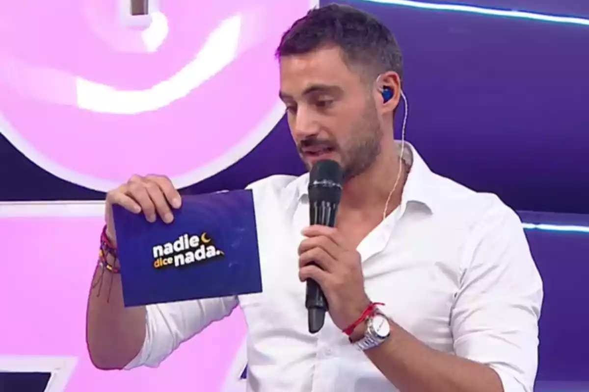 A man in a white jersey holds a microphone and a card that says "nobody says anything" on a television set with a pink background.